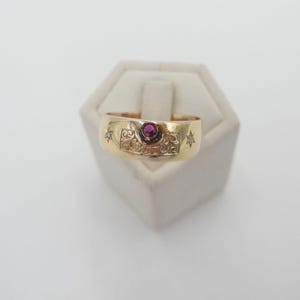 May include: A gold band ring with a red ruby stone and small diamonds set in a star shape. The ring has an intricate design.