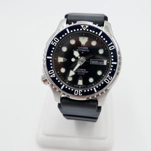 Könnte beinhalten: Eine schwarze Citizen Automatik-Taucheruhr mit schwarzem Kautschukband. Das Zifferblatt der Uhr hat weiße Markierungen und ein Datumsfenster mit der Aufschrift "MON 12". Die Uhr ist wasserdicht bis 200 Meter.