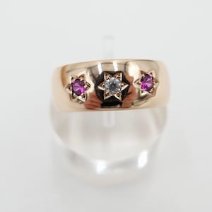 Op de afbeelding: Een gouden ring met drie stenen die in stervormige settings zijn gezet. De middelste steen is een heldere diamant en de twee buitenste stenen zijn roze.