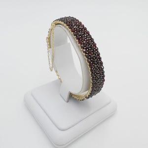 May include: A gold bangle bracelet with a row of red garnet stones. The bracelet is on a white display stand.