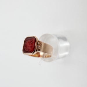 Puede incluir: Un anillo de oro con una piedra preciosa roja rectangular con un diseño tallado. El anillo tiene una banda detallada con un patrón adornado en los lados. La piedra preciosa está engastada en un bisel de oro.