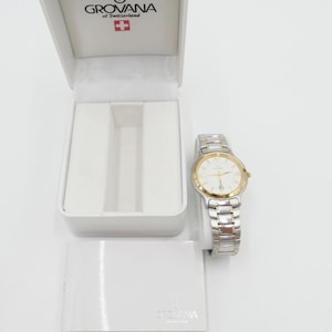 Könnte beinhalten: Eine weiße und goldene Grovana-Uhr mit weißem Zifferblatt und silbernen Zeigern. Die Uhr befindet sich in einer weißen Box mit dem Grovana-Logo und den Worten "of Switzerland".