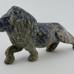 Puede incluir: Una escultura de león de piedra en posición de caminar. El león tiene una superficie texturizada con una mezcla de colores azul, gris y beige. La escultura está sobre un fondo blanco.