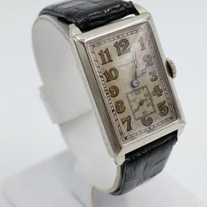 Puede incluir: Un reloj de pulsera Longines de plata con caja rectangular y correa de cuero negro. La esfera del reloj tiene marcadores de hora en números romanos y un pequeño dial de segundos.