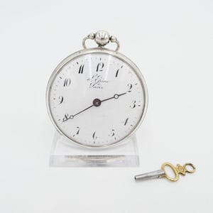 May include: A silver pocket watch with a white face and black Roman numeral hour markers. The watch has a key wind mechanism and is displayed on a clear stand. The watch face has the text "Lejeune Paris" printed on it.