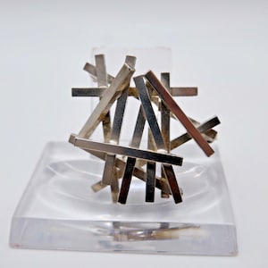May include: A silver-toned brooch composed of multiple intersecting rectangular metal bars arranged in a chaotic, layered grid pattern. The metallic surface reflects light, creating varying shades of silver and grey across the geometric structure.