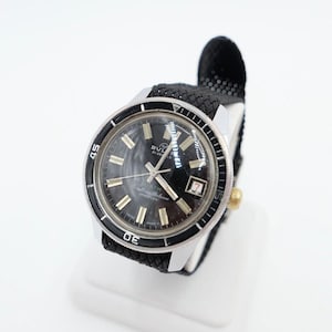 Könnte beinhalten: Eine schwarz-silberne Armbanduhr mit schwarzem Zifferblatt und schwarzem Nylonarmband. Die Uhr hat ein Datumsfenster bei 3 Uhr und eine drehbare Lünette.