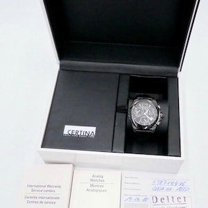 Könnte beinhalten: Eine schwarze Uhr mit silbernem Zifferblatt und Chronographenfunktionen. Die Uhr befindet sich in einer weißen Box mit dem Markennamen "Certina" darauf. Die Box enthält auch eine Garantiekarte und eine Bedienungsanleitung.