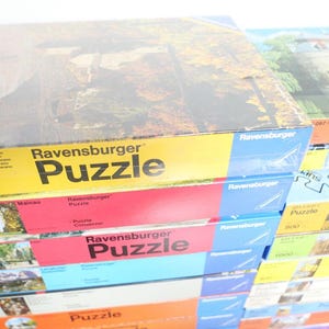 May include: A collection of Ravensburger jigsaw puzzle boxes. The boxes are in various colours, including yellow, red, and blue. The word "Puzzle" is clearly visible on each box. The puzzles seem to depict landscape scenes.