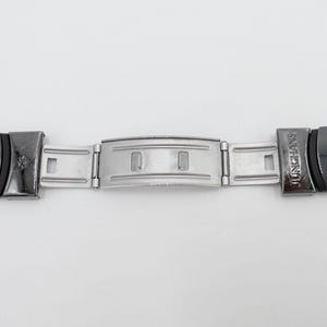 Könnte beinhalten: Ein silbernes Uhrenarmband mit schwarzen Endstücken. Die Endstücke sind mit dem Wort "JUNGHANS" gekennzeichnet.