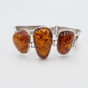 Könnte beinhalten: Ein silbernes Armband mit drei Bernstein-Steinen. Die Steine sind oval geformt und haben eine braune Farbe.