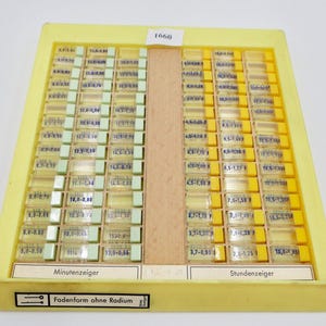 May include: A yellow plastic tray with two rows of small glass vials containing watch hands. The vials are labeled with numbers and measurements in millimeters. The tray is labeled "Minutenzeiger" and "Stundenzeiger" for minute and hour hands, respectively. The tray also has a label that reads "Fadenform ohne Radium" which translates to "Thread form without radium".