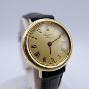 Könnte beinhalten: Eine goldfarbene Chopard Genève Armbanduhr mit schwarzem Lederarmband. Das Zifferblatt der Uhr ist goldfarben mit schwarzen römischen Ziffern und Zeigern. Die Wörter "Chopard Genève" und "Swiss Made" sind auf dem Zifferblatt aufgedruckt.