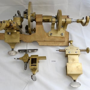 Op de afbeelding: Een vintage draaibank van messing met een houten basis en twee extra hulpstukken. De draaibank heeft een handwiel en een tandwielsysteem. De hulpstukken zijn ook van messing en hebben een vergelijkbaar ontwerp.