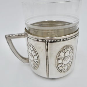 May include: Silver-plated glass holder with a floral design. The holder has a beaded rim and a handle.