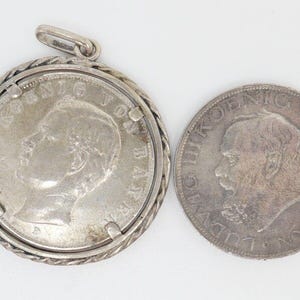 May include: A silver coin pendant with a portrait of a man on one side and a design on the other. The pendant is attached to a silver chain.