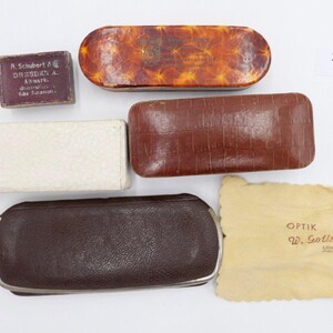 May include: Five vintage eyeglass cases in various colors and materials, including brown leather, white cardboard, and a brown cloth cleaning cloth with the text "OPTIK W. Gottscheich" printed on it. The number "2326" is written on a white label.