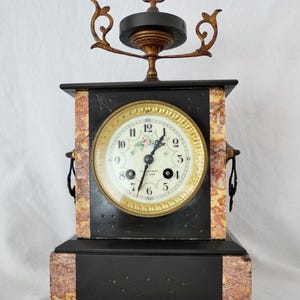 Könnte beinhalten: Eine schwarze und goldene Kaminuhr mit einem Marmorsockel und einem dekorativen Aufsatz. Das Zifferblatt der Uhr hat römische Ziffern und ein Blumenmuster.