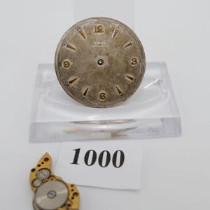 May include: A silver watch face with black numbers and hands. The watch face has the text "EDEL" and "MATIC" printed on it. The watch face is on a clear plastic stand. There is a small watch part on a white surface with the number "1000" printed on it.
