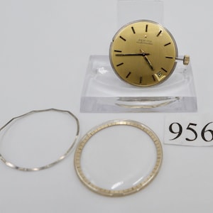 May include: A gold-coloured Zenith automatic watch face with black markers and hands, and a date window at the 3 o'clock position. The watch face is displayed on a clear acrylic stand, with a silver bezel and a clear watch crystal. The number "956" is on a white paper.