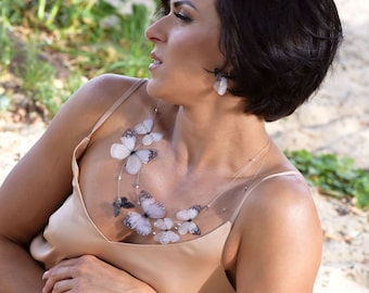 Op de afbeelding: Een vrouw met kort donker haar draagt een beige tanktop met een delicate ketting met meerdere witte vlinderbedels.