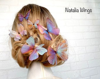 May include: A woman's hair is adorned with multiple butterfly hair clips. The butterflies are made of iridescent fabric and have a purple, pink, and blue color scheme. The hair clips are arranged in a cluster on the side of her head. The text "Natalia Wings" is visible in the upper right corner of the image.