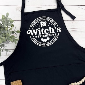 May include: Black kitchen apron with a white circle design that says "Premium Witch's Brew Witch's Kitchen Stirring Up Some Magic".