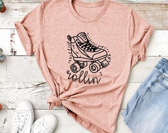 May include: A light pink t-shirt featuring a black and white illustration of a roller skate with the word "rollin'" in a script font. The shirt is paired with blue jeans and white sneakers.