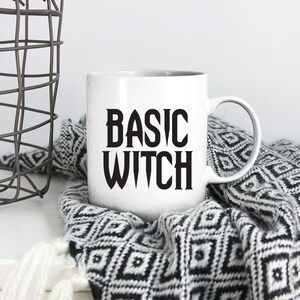 May include: White ceramic mug with black text that reads "BASIC WITCH" on a white surface with a black and white patterned blanket.
