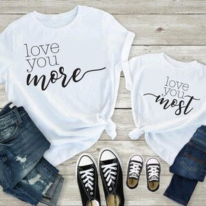 May include: Two white t-shirts with black text. The larger shirt says "love you more" and the smaller shirt says "love you most". The shirts are laid out on a wooden surface with blue jeans and black sneakers.