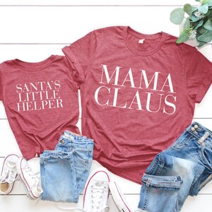 May include: Two red t-shirts with white text. The larger shirt says "Mama Claus" and the smaller shirt says "Santa's Little Helper".