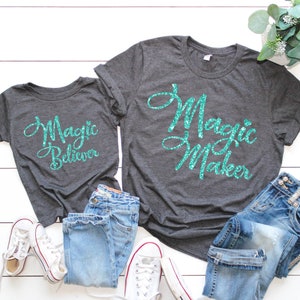 May include: Two gray t-shirts with teal glitter text. The larger shirt says "Magic Maker" and the smaller shirt says "Magic Believer".