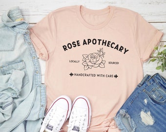 Può includere: Una maglietta rosa con il testo "ROSE APOTHECARY" in lettere nere. Il testo è arcuato sopra un disegno a linea nera di una rosa. Sotto la rosa, il testo recita "LOCALLY SOURCED" e "HANDCRAFTED WITH CARE".