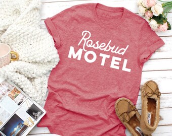 May include: A red t-shirt with the text "Rosebud Motel" in white lettering. The shirt is on a white wooden surface with a white blanket, a magazine, and a pair of brown moccasins.