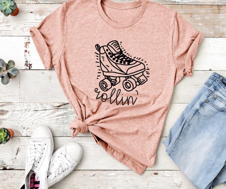 May include: A light pink t-shirt featuring a black and white illustration of a roller skate with the word "rollin'" in a script font. The shirt is paired with blue jeans and white sneakers.