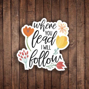 May include: A white sticker with the text "where you lead I will follow" in black script. The sticker is decorated with orange, yellow, and red floral illustrations. The background is a wooden surface.