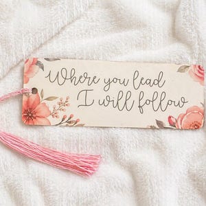 May include: A rectangular bookmark with the text "Where you lead I will follow" in elegant script. The bookmark features floral watercolor designs in shades of pink and green, and a pink tassel. The background is a white, textured surface.
