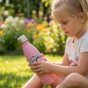 May include: A light pink stainless steel water bottle with a silver cap. The bottle features the name Sophia printed in black script lettering, accompanied by a small red bow graphic positioned above the letter i in the name.