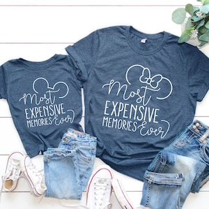 May include: Two navy blue t-shirts with white text that reads "Most Expensive Memories Ever" and a white outline of Mickey Mouse ears. The larger shirt has a white bow on the ears. The shirts are laid out on a white wooden surface with blue jeans and white sneakers.
