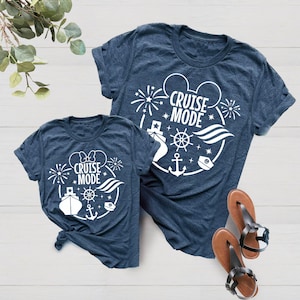 Pode incluir: Duas camisetas azul marinho com um contorno branco das orelhas do Mickey Mouse, um navio, uma âncora, um volante e fogos de artifício. O texto "Cruise Mode" está dentro das orelhas do Mickey Mouse.