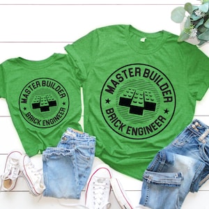 May include: Two green t-shirts with a black circle design that says "Master Builder Brick Engineer" and a picture of a Lego brick. The shirts are on a white wooden surface with blue jeans and white sneakers.