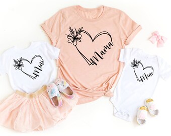 Könnte beinhalten: Zwei weiße T-Shirts mit schwarzen floralen Herzmotiven. Das größere Shirt sagt "Mama" und das kleinere Shirt sagt "Mini".
