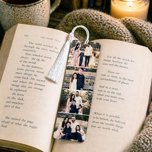 May include: A personalized bookmark featuring family photos, with a white tassel, rests inside an open book. A cup of tea and a lit candle are in the background, creating a cozy reading scene. The bookmark is a sentimental gift.