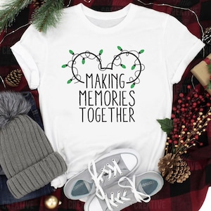 May include: White t-shirt with the text "MAKING MEMORIES TOGETHER" in black. The design includes a Mickey Mouse head shape made of Christmas lights. The shirt is paired with gray sneakers, a gray beanie, and a gift box.
