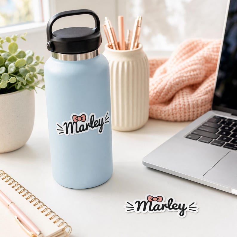 May include: A light blue water bottle with a black lid and a white sticker featuring the name Marley in black cursive script, accented by a small pink bow and whiskers. A matching sticker is placed on the surface next to the bottle.