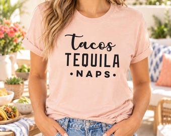 Puede incluir: Una camiseta de manga corta de color rosa claro con el texto negro "Tacos TEQUILA • NAPS •" impreso en la parte delantera. La camiseta tiene cuello redondo, mangas remangadas y se combina con vaqueros azules.