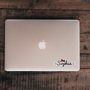 May include: A silver laptop with a white apple logo in the center and a custom sticker applied to the lower right corner. The sticker features the name Sophia in black cursive script, accented by a small red bow and short black lines on each side.
