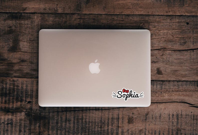 May include: A silver laptop with a white apple logo in the center and a custom sticker applied to the lower right corner. The sticker features the name Sophia in black cursive script, accented by a small red bow and short black lines on each side.