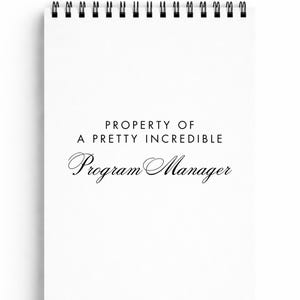 May include: A white spiral-bound notepad with black metal rings at the top. The text "PROPERTY OF A PRETTY INCREDIBLE Program Manager" is printed in black on the front of the notepad.