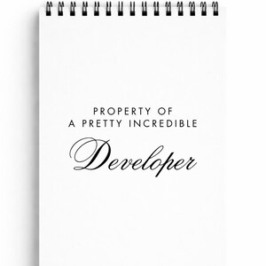May include: White spiral-bound notepad with black metal rings. The cover features the text "PROPERTY OF A PRETTY INCREDIBLE Developer" in black script and sans-serif fonts. The notepad is blank inside, ready for notes.